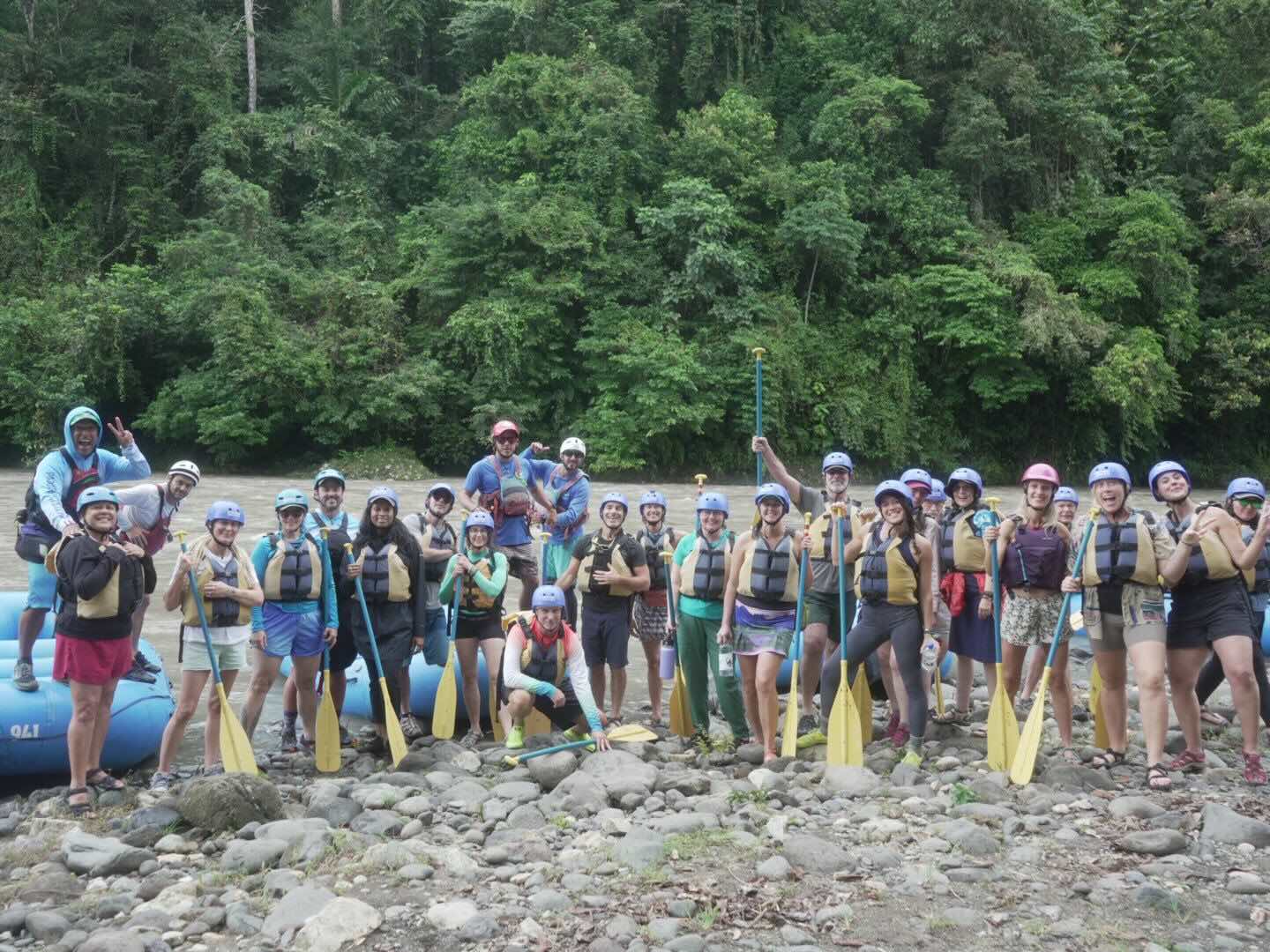 Group of 25 people standing along river bank holding paddles and wearing rafting gear